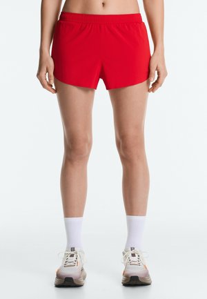 Person iført røde atletikshorts, hvide crew-sokker og hvide løbesko står mod en ensfarvet baggrund.
