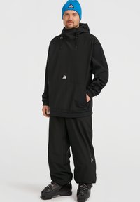 Zwarte pullover met een capuchon en een voorzak, voorzien van een driehoekig logo; gecombineerd met loszittende zwarte broek en zwarte laarzen. Blauwe gebreide muts.