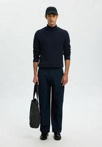 Jauns vīrietis, tērpies tumši zilā turtlenecka džemperī ar atbilstošām biksēm, melnā cepurē un zābakiem, tur tumšu iepirkumu maisu uz vienkārša fona.