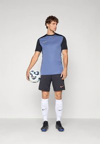 Fiatal férfi kék-fekete sportöltözetben, kezében futballlabdával, fehér zoknit és fekete stoplis cipőt viselve, sima háttér előtt áll.