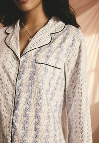 Valkoinen pyjaman paita, jossa on sininen kukkakuosi, mustat reunat ja pieni tasku. Ominaisuuksina pystysuorat viivakuviot ja nappikiinnitys.