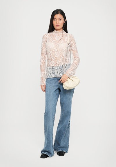 Kanten top met lange mouwen en bloempatronen, gecombineerd met lichtblauwe flared jeans. Model houdt een crèmekleurige pouch tas vast.