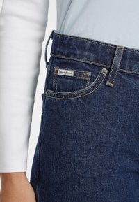 Jeans de mezclilla azul oscuro con un corte recto, que presentan un botón plateado y costuras amarillas, además de una etiqueta "Guess Jeans" en el bolsillo delantero.