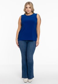 Blå ærmeløs top i glat stof, rund halsudskæring, parret med klassiske klokkeformede denimjeans og hvide sko. Simpelt og tætsiddende design.