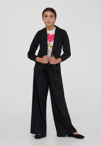 Blazer nero con maniche lunghe e risvolti, abbinato a pantaloni neri a gamba larga e a una t-shirt bianca con un design di cuore rosa.