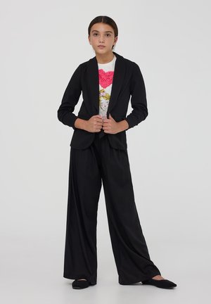 Blazer nero con maniche lunghe e risvolti, abbinato a pantaloni neri a gamba larga e a una t-shirt bianca con un design di cuore rosa.