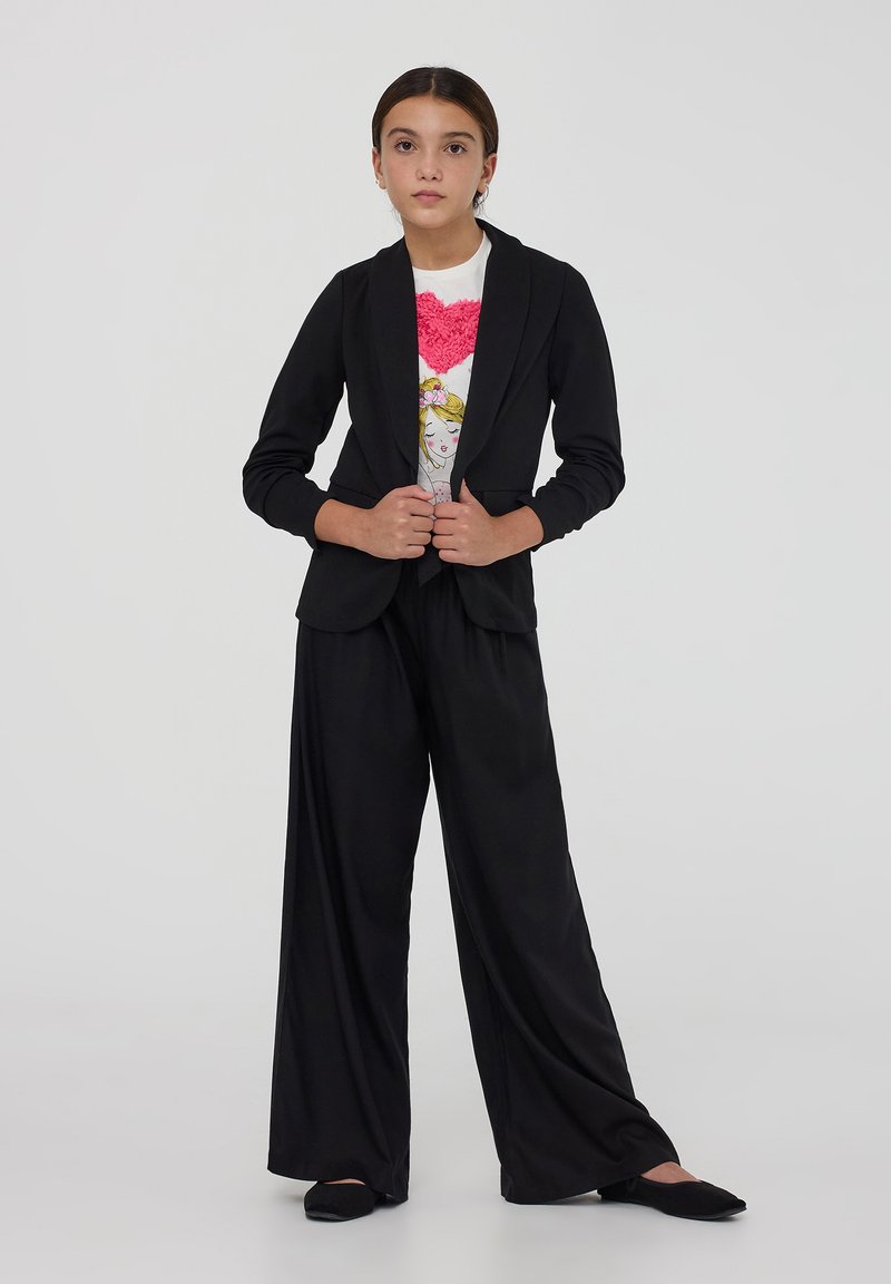 Blazer nero con maniche lunghe e risvolti, abbinato a pantaloni neri a gamba larga e a una t-shirt bianca con un design di cuore rosa.