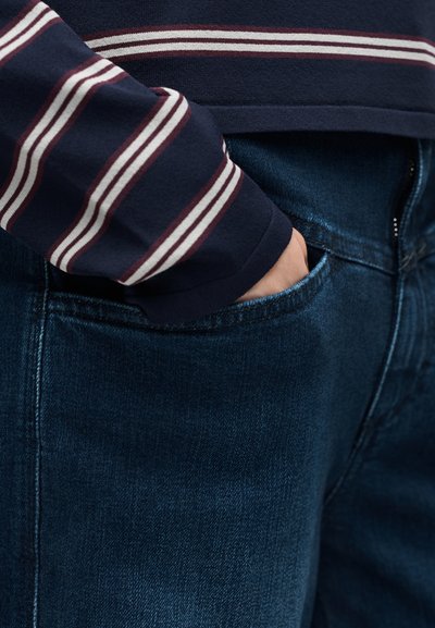 Χέρι στην μπροστινή τσέπη σκούρου μπλε τζιν, που φορεί μπλε σκούρο πουλόβερ με λευκές και κόκκινες οριζόντιες ρίγες στο μανίκι.