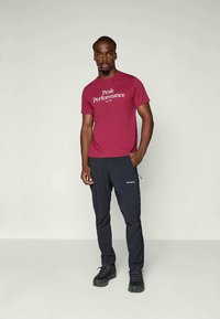 Červené tričko s nápisem "Peak Performance", v kombinaci s tmavě modrými kalhotami a černými turistickými botami. Hladký materiál, volný střih a funkční design.