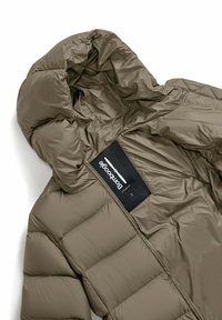 Pufferjack in taupe met een glanzende afwerking, voorzien van een capuchon, horizontaal gestikt patroon en een zwart label met witte tekst aan de binnenkant.