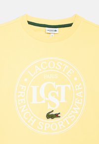 Keltainen puuvillainen collegepaita, jossa on pyöreä valkoinen logo, jossa lukee "LACOSTE PARIS FRENCH SPORTSWEAR" ja vihreä kirjottu krokotiilikoriste.
