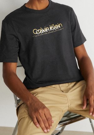 Zwarte katoenen T-shirt met een opvallend gouden "Calvin Klein" logo over de borst. Korte mouwen en ronde hals. Soepele textuur.