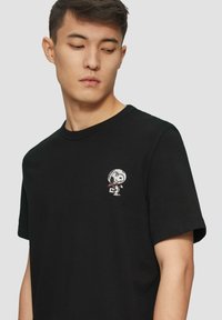 Czarna bawełniana koszulka z krótkim rękawem, z małym haftowanym wzorem Snoopy po lewej stronie klatki piersiowej. Okrągły dekolt, luźny krój.