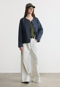 Γυναίκα που φοράει ναυτικό μπουφάν με καπιτονέ, πράσινο τοπ, λευκό παντελόνι με φαρδιά πόδια, μαύρα παπούτσια και καρό καπέλο μπέιζμπολ, στέκεται μπροστά σε λευκό φόντο.