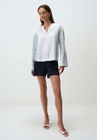 Camicia leggera, con bottoni, di colore bianco con maniche blu, caratterizzata da un fit rilassato e scollatura a V. Abbinata a pantaloni corti blu navy e tacchi bianchi.