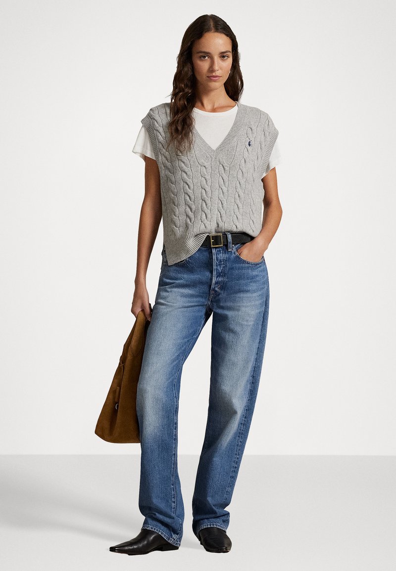 Grå kabelstrikket ærmeløs sweater over en hvid T-shirt, parret med lyseblå straight-leg jeans og sorte spidse støvler. Brun taske.