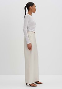 White long-sleeve top paired with creamy wide-leg trousers, featuring a relaxed fit. The model wears dark sandals; background is plain.