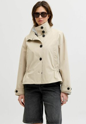 Femme portant une veste beige boutonnée avec un col montant, des lunettes de soleil noires et un short en jean noir, debout devant un fond uni.