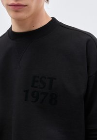 Νεαρό άτομο που φοράει μαύρο φούτερ με στρογγυλή λαιμόκοψη και ανάγλυφ ο κείμενο "EST 1978" στο στήθος, μπροστά σε απλό φόντο.