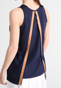 Marinblå ärmlös topp i texturerat tyg, med ett centralt beige rand och små perforeringar för ventilation på baksidan.