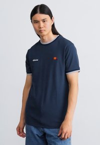 Marineblå bomulds T-shirt med hvid ribbet rund hals og detaljer i ærmerne, med et lille rødt og orange logo på venstre bryst. Modellen bærer blå jeans.