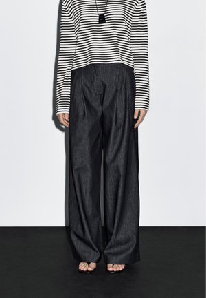 Zwarte wijde spijkerbroek met plooien, gecombineerd met een cropped zwart-wit gestreept langemouwen top en simpele sandalen.