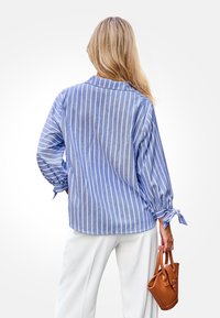 Gestreiftes langärmliges Hemd in Blau und Weiß mit Manschetten zum Binden, kombiniert mit weißen Hosen und einer kleinen braunen Handtasche. Glatte Textur.