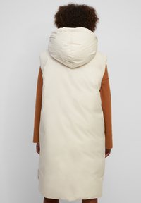 Manteau sans manches de couleur crème, rembourré, avec une grande capuche, texture lisse et étiquette sur le côté. La vue arrière montre un design jusqu'aux genoux. Pull marron en dessous.