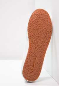 Weiße Sneaker mit einer strukturierten Gummisohle und braunen Akzenten. Das Obermaterial ist glatt mit minimalen Designdetails. Markennamen auf der Sohle eingestanzt.