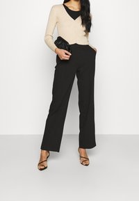 Kvinna som bär en beige knäknappad kofta över en svart topp, svarta vida byxor, svarta sandaletter med remmar, och håller en svart läderclutch.