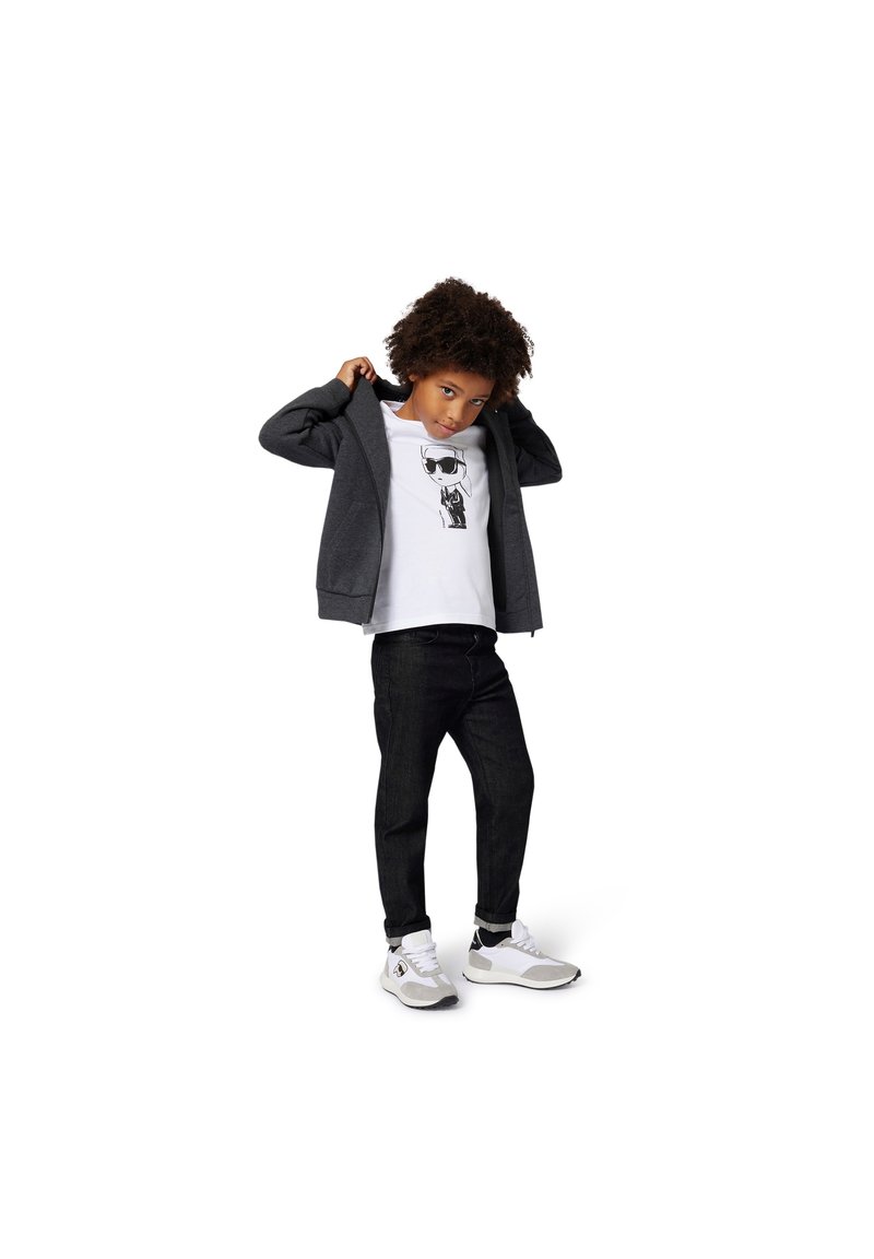 Šedá mikina na zip přes bílé tričko s grafickým motivem, doplněná tmavými džíny a bílými teniskami s černými detaily.