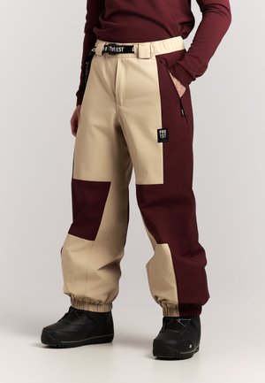 Tweekleurige beige en bordeauxrode broek met elastische boorden, een zwarte riem, zijritszakje en zwarte schoenen bij een persoon die een bordeauxrode top draagt.