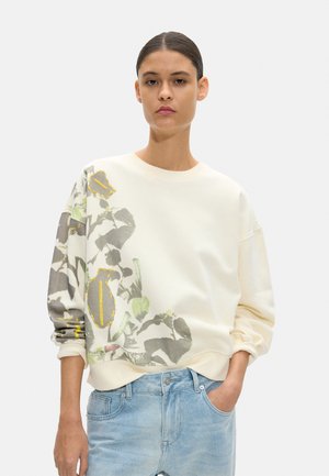 Kobieta ubrana w kremową bluzę z szaro-zielonym botanicznym wzorem i żółtymi szwami, w połączeniu z jasnoniebieskimi dżinsami, pozująca z jedną ręką w kieszeni.