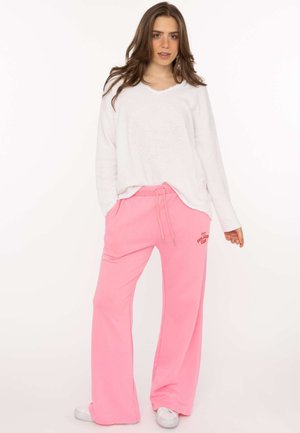 Frau trägt ein weißes Langarmshirt und pinke weite Jogginghosen mit dem Text „Feel Good Club“, posiert mit Händen in den Taschen und weißen Sneakers.