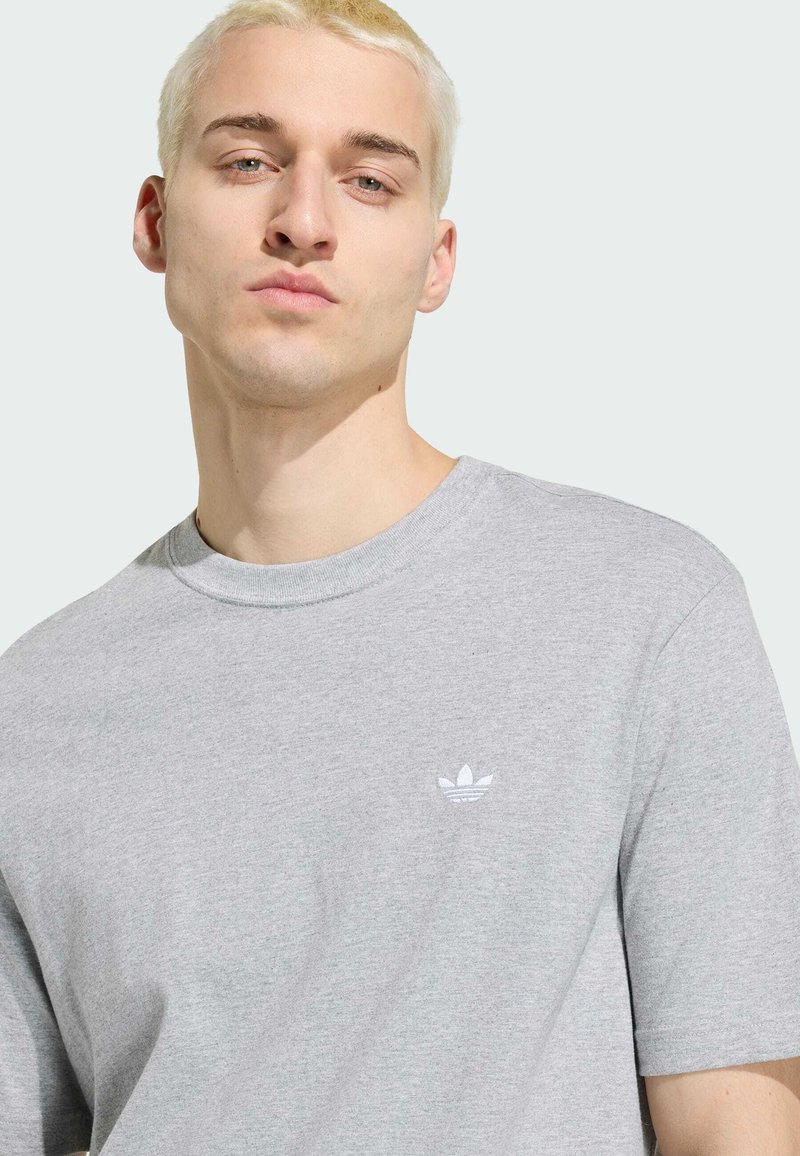 Ung mand med kort blondt hår iført en lysegrå Adidas t-shirt med et lille hvidt logo på brystet, neutral baggrund.