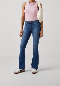 Mujer con una blusa sin mangas rosa, jeans acampanados azules, zapatillas blancas, llevando un bolso blanco en un fondo gris claro.