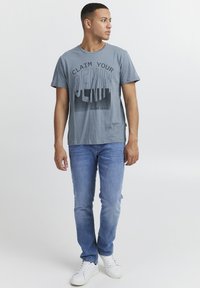 Grå t-skjorte med "CLAIM YOUR DENIM" grafikk trykket i svart. Kombinert med lyseblå jeans og hvite joggesko. Uformell design, kortermet.