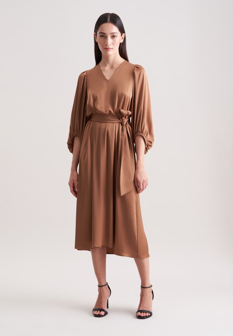 Braunes, knielanges Kleid mit V-Ausschnitt, gepufften Ärmeln und einem Taillengürtel. Hergestellt aus glattem Stoff mit fließendem Design und minimalistischer Ästhetik.