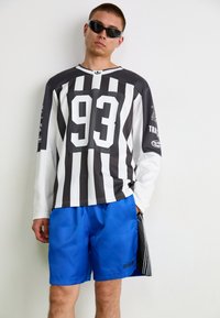 Čierno-biele pruhované dlhé tričko s veľkým "93" na prednej strane, skombinované s modrými šortkami s čiernymi akcentmi a logom Adidas. Nosené slnečné okuliare.