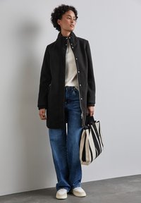 Svart ullkappa med hög krage, silverfärgade tryckknappar, ihop med en krämfärgad tröja och vida blå jeans. Håller en svart och vit randig tote-väska.