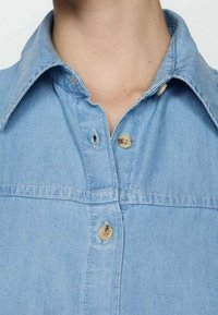 Close-up of a person wearing a light blue denim shirt with the top two buttons undone, showing the neck and collar area.