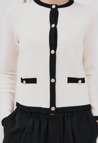 Cardigan lavorato a maglia di colore crema con bordo a contrasto nero e cinque bottoni decorativi. Presenta due tasche nere con dettagli dorati. Texture morbida.