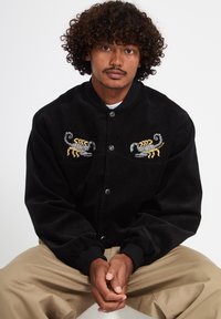 Homme aux cheveux bouclés portant une veste noire brodée de deux scorpions jaunes et gris, assis les mains jointes sur un pantalon beige.