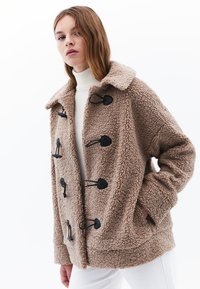 Ruskea fleece-takki, jossa on teksturoitu pinta, kaulus, mustat napit ja sivutaskut. Päällä valkoinen toppi.