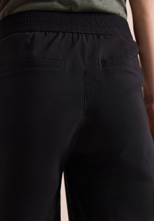 Lähikuva mustista housuista, joissa on joustava vyötärö ja kaksi takana olevaa piilotettua taskua, yllään vihreä toppi.