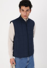Marineblauer gesteppter Weste mit hohem Kragen und Reißverschluss, getragen über einem leichten Pullover; verfügt über ein quadratisches Muster und eine glatte Textur.