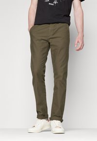 Oliwkowo-zielone bawełniane spodnie o prostym kroju, z bocznymi kieszeniami i mankietami, noszone z białymi trampkami i czarnym T-shirtem z nadrukiem.