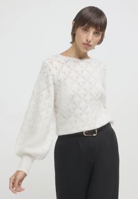 Weißer Pullover mit Rautenmuster, runder Ausschnitt und Ballonärmeln, kombiniert mit hoch geschnittenen schwarzen Hosen und einer goldenen Gürtelschnalle.