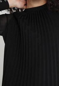 Blusa nera plissettata con scollo alto e maniche lunghe trasparenti. Presenta plissettature verticali e una leggera lucentezza, che offrono texture e profondità.
