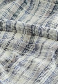 Tissu à carreaux en blanc, marine et gris, avec une finition texturée. Présente un bouton rond de couleur claire au milieu des plis.